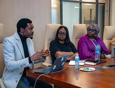 Three people in a boardroom: a man in a light-blue blazer speaks at a laptop with two women nearby.