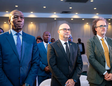 Photograph of three men in suits standing at a conference, with an audience behind.