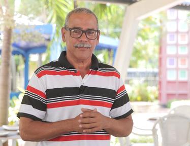 Smiling man with glasses in a striped polo stands outdoors under a pavilion with hands clasped.