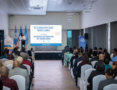 Conference hall with seated audience facing stage; presenter at podium near projection screen.