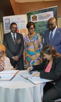 Photo: Two people sign documents at round table while four colleagues stand in a conference room.