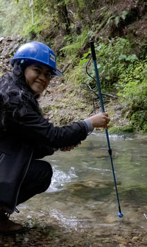 Photograph of a person wearing a blue helmet kneeling by a shallow stream, holding a long pole.