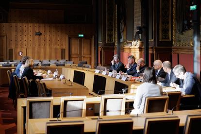 A formal meeting in a grand room with attendees seated at a long table.