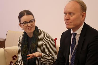 A woman in glasses smiles and gestures while speaking to a man in a suit.