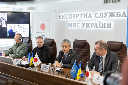 Conference setting with four men seated at a table, flags displayed, discussing a topic.