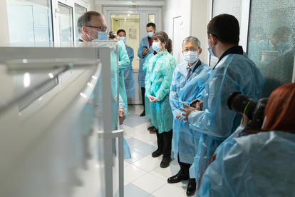 Group of people in protective gear and masks, listening to a speaker in a hospital corridor.