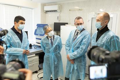 Four men in blue protective gowns and masks discuss in a lab setting, surrounded by cameras.