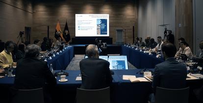 A conference room filled with people seated around a long table and a presentation on screen.