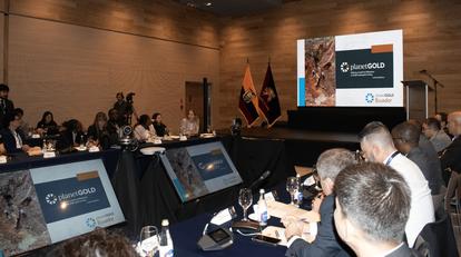 A conference room with attendees sitting at a table, a presentation screen in the background.