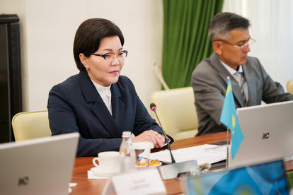 A woman in a suit sits at a conference table, focused, with a flag and laptops in the background.
