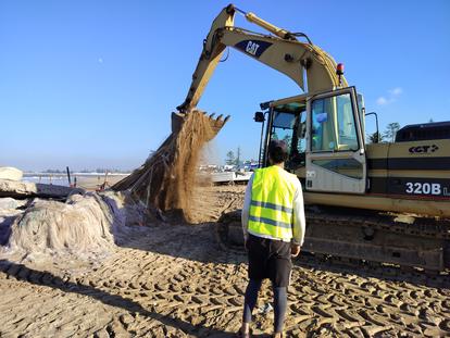 Excavator lifting sand on a beach, with a person in a yellow safety vest observing.