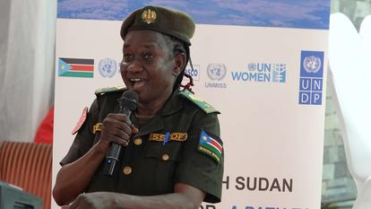 A smiling woman in military uniform speaks into a microphone at an event backdrop.
