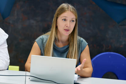 A woman with long hair gestures while speaking at a table with a laptop and notepad.