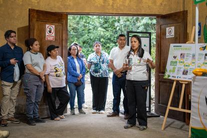 A group of people gathered indoors, listening to a woman speaking with a presentation board.
