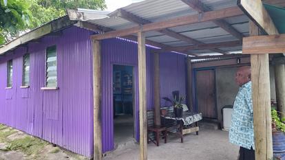 A purple building with a covered porch, featuring a man standing nearby and green plants.