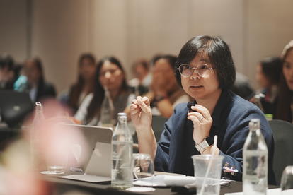 A woman with glasses attentively participates in a conference, surrounded by an audience.
