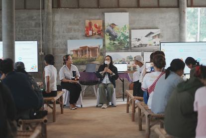 A woman speaking to an audience in a rustic hall, with artwork on the walls and attendees seated.