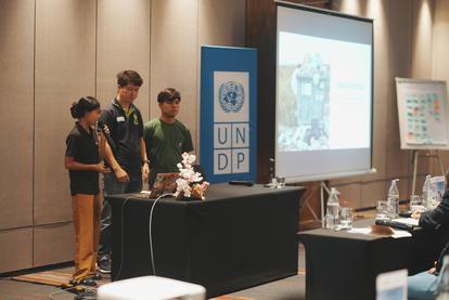 Three individuals present at a conference table with a UNDP banner in the background.