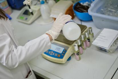 A gloved hand pours a liquid from a bottle onto a scale in a lab setting.