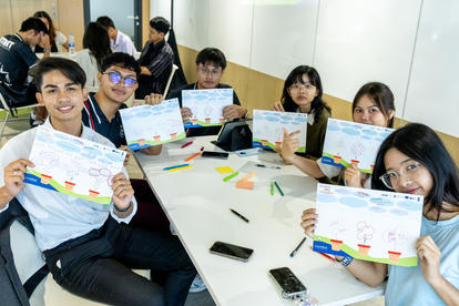A group of six young people smile and hold colorful drawings at a work table.