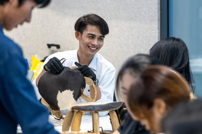 A smiling man in gloves works on a sculpture while others observe in a workshop setting.