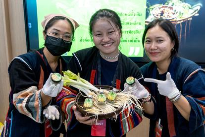 Three women in traditional attire smile and hold a tray of colorful dishes, with a vibrant background.