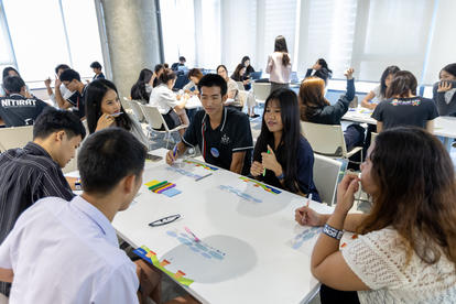 A group of young adults engaged in discussion around a table with colorful materials in a bright room.