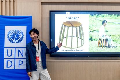 A person gestures towards a presentation of a wooden stool next to a UNDP banner.