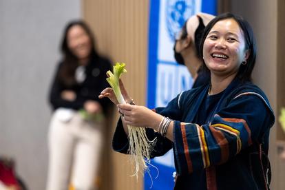 Woman in traditional clothing smiling while holding fresh vegetables, with a blurred figure in the background.