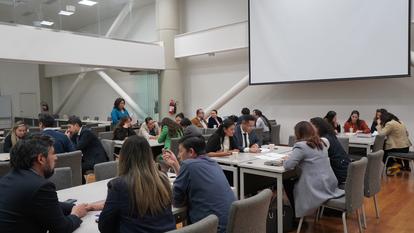 Participants around tables in a conference room, some raising hands, projector screen at front.