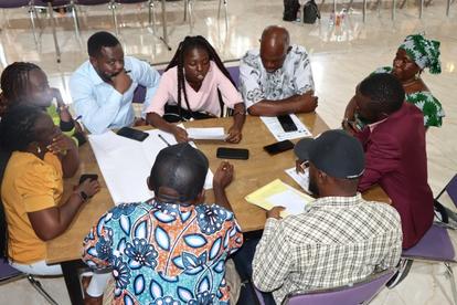 Group of adults around a circular table, examining documents in a bright room.