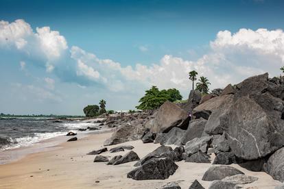 Sunny beach with large rocks along the shore, palm trees, and a blue sky with clouds.