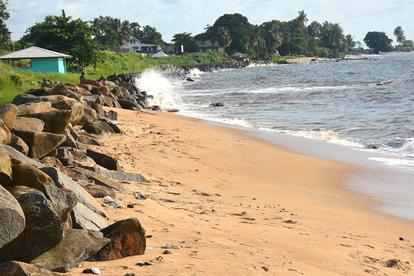 Sunny beach with rocky shoreline on the left, golden sand and gentle waves; green houses in back.