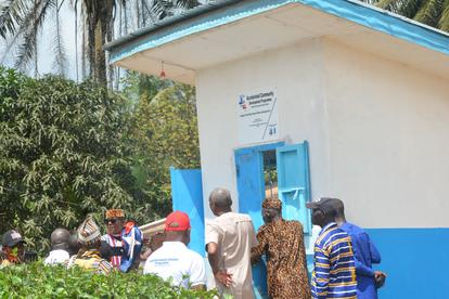 Photograph of a small cream building with blue doors and trim, a crowd gathered outside.
