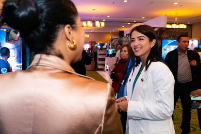 Person in a metallic copper jacket talks to a woman in a white lab coat at a crowded indoor event.