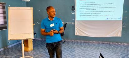 Photograph: Smiling speaker in a blue polo presenting in a turquoise room with projector screen.