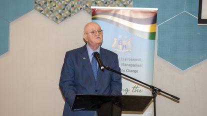 Photograph of an older man in a blue suit speaking at a lectern, banner behind him.