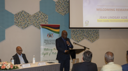 Man at podium delivering remarks at a conference; panelists at table with flowers, screen in background.