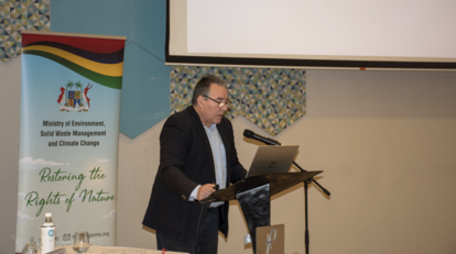 A man in a dark suit speaks at a podium beside a rainbow banner; projector screen behind.