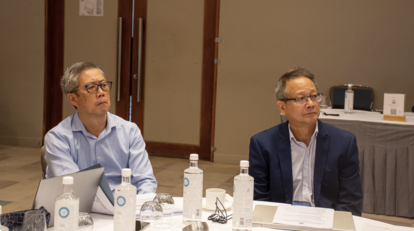 Two men in a conference room sit at a table with papers, water bottles, and a laptop.