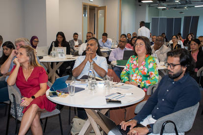 Photograph of diverse conference attendees seated around round tables, listening attentively.