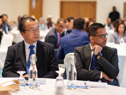 Two men in suits sit at a conference table with water bottles and papers, surrounded by attendees.