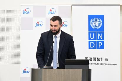 Man in suit speaks at a podium with UNDP and JICA banners.