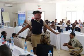 Woman wearing a mask demonstrates with arms extended at a classroom workshop; attendees at tables.