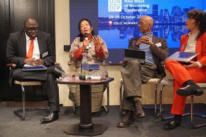 Panel discussion with four speakers seated in a row on stage; blue backdrop with UN branding.