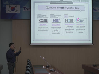 Presenter gestures toward a projector slide displaying Korea statistics services logos in a conference room.