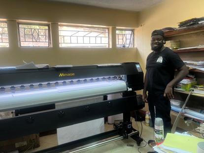 Photograph of a man standing beside a large-format printer in a yellow-walled workshop.
