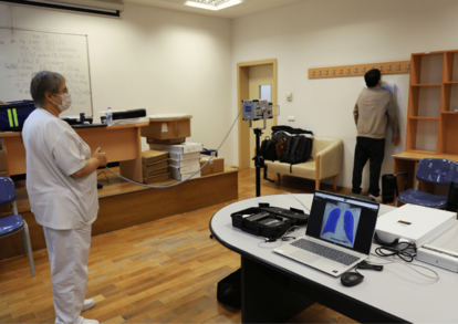 Two people in a computer lab: one in white coat at a desk with laptops, another near shelves.
