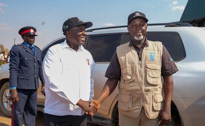 Three men in uniform and vest shake hands beside a small silver airplane.