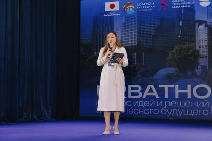 Female presenter in a white dress speaks into a microphone on a blue-lit stage.